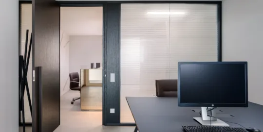Ufficio moderno con scrivania, monitor e porta scorrevole in vetro, arredamento minimalista e colori neutri. La lampada sopra la scrivanie è a basso abbagliamento con diffusore a nido d'ape.