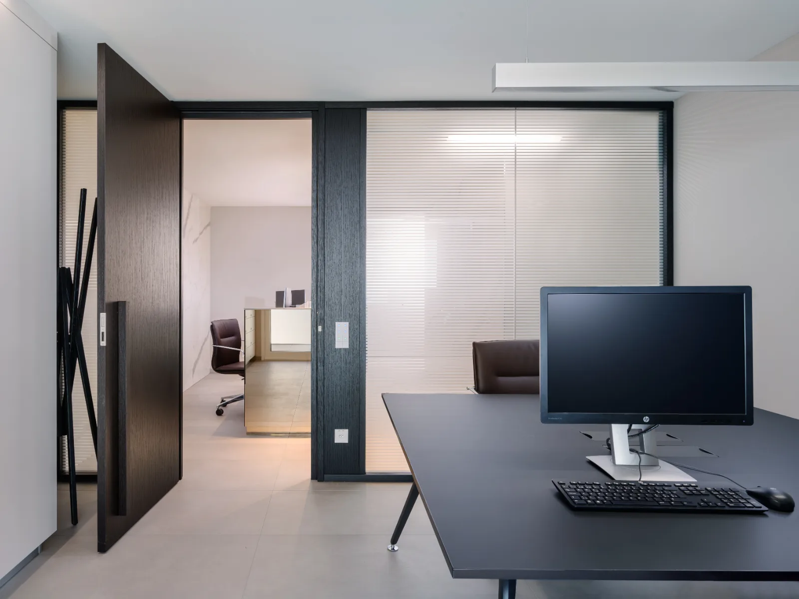 Ufficio moderno con scrivania, monitor e porta scorrevole in vetro, arredamento minimalista e colori neutri. La lampada sopra la scrivanie è a basso abbagliamento con diffusore a nido d'ape.