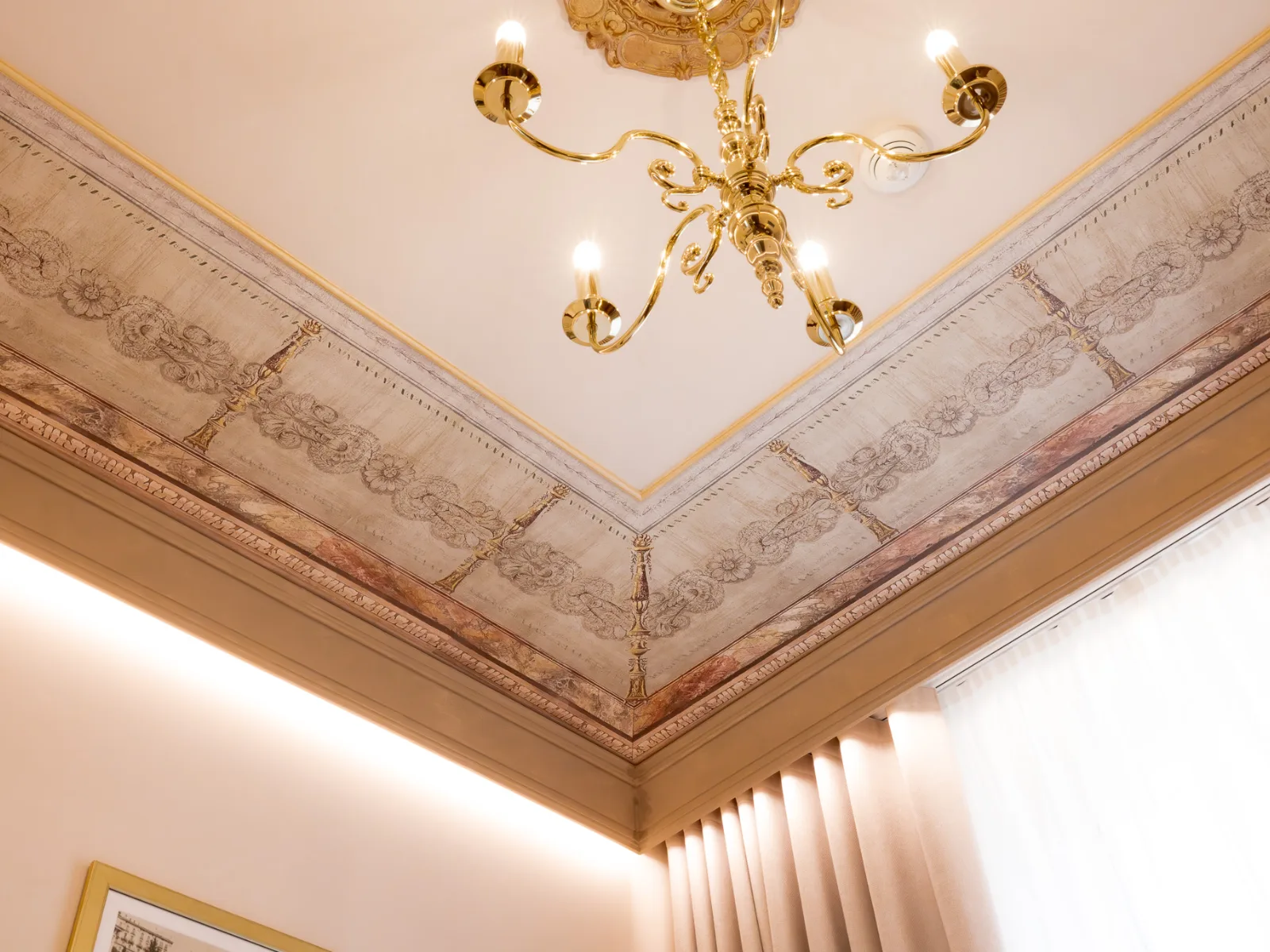 Dettaglio di un soffitto decorato con motivi floreali e un elegante lampadario dorato. Si vede una Veletta che copre la tenda ed illumina il perimetro